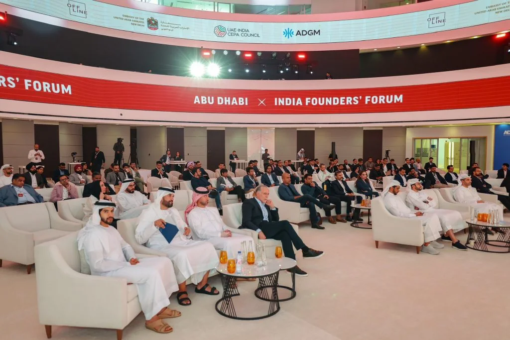The venue in the round: the audience in white armchairs beneath a curved red LED ring reading “Abu Dhabi × India Founders’ Forum”, with partner logos above.