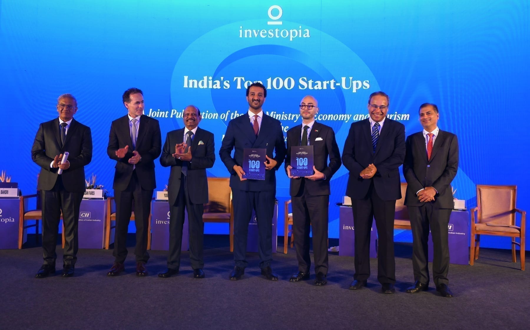Seven officials on stage at Investopia Global Ahmedabad, two holding up copies of the dossier, in front of a blue backdrop reading “India’s Top 100 Start-Ups”.